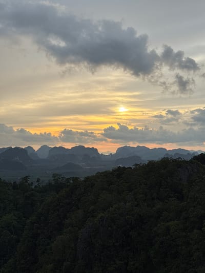 This is a landscape photograph taken in the late afternoon, capturing a view from a high vantage point overlooking a lush, forested valley and a series of karst mountains in the distance. The sky is filled with a dramatic sunset, with streaks of orange and yellow light breaking through scattered grey clouds. The foreground is dominated by a dense, dark green forest, with trees and foliage creating a textured, almost silhouetted appearance. Beyond the immediate forest, the landscape opens up to reveal a valley with scattered vegetation and more distant, jagged mountain peaks that are silhouetted against the colorful sky. The overall impression is one of natural beauty and tranquility, with the setting sun casting a warm glow over the scene.