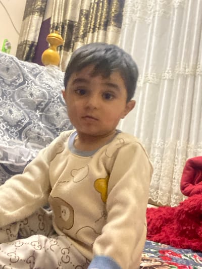 An indoor scene in Galaosiyo, Uzbekistan, depicts a young child seated and looking directly at the viewer. The child, with dark hair and eyes, wears cream-colored long-sleeved pajamas featuring blue trim at the neck and cuffs, and cartoon-style graphics including outlines of clouds, a yellow shape, and a brown bear. The lower garment appears to be light-colored patterned pants with a repeating "GG" logo. The background on the left includes a dark grey or blue patterned fabric with white designs, and a dark patterned curtain with vertical gold and brown motifs. A yellow, hourglass-shaped decorative object rests on a surface behind the child's left shoulder. To the right, a light-colored sheer curtain with a lace-like pattern is visible, alongside a segment of vibrant red, textured fabric, possibly a blanket or plush item.