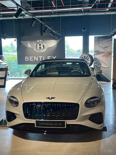 A cream-colored Bentley Continental GTC V8 convertible is parked indoors, facing the camera. The car has a black grille and a black convertible top. The Bentley logo is visible on the front of the car. Behind the car, a dark wall features the Bentley logo and the word "BENTLEY" in large white letters. The environment appears to be a car showroom, with track lighting overhead and large windows in the background showing trees and greenery. There is a spare tire on display behind the car, propped up vertically. A smaller wheel is visible to the right. The floor is polished concrete, reflecting the car. A sign below the grille reads "CONTINENTAL GTC V8". The setting is the Bentley dealership in Reckange-sur-Mess, Luxembourg. There are no people visible in the image.