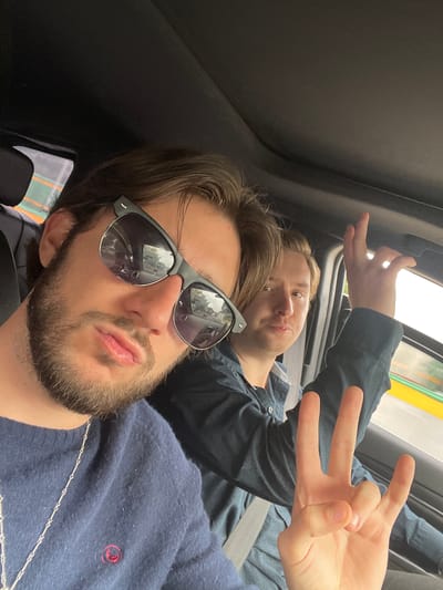 Two individuals are in a vehicle, likely a car, with one in the foreground and the other behind. The person in the foreground is a young man with light brown hair, a beard, and sunglasses. He is wearing a dark blue sweater with a small red logo on the left chest and a silver necklace. He is looking at the camera with his lips pursed. The person behind him, also a young man with light brown hair, is wearing a dark blue collared shirt and has his right hand raised, forming a peace sign with two fingers extended and the other two tucked in, while his thumb is extended, making a 'rock on' gesture. The background shows a view through the car window of an urban environment with blurred buildings and yellow and green lines on what appears to be a road or pedestrian walkway. The interior of the car is visible, including the black headliner and a portion of a seat. The lighting suggests it is daytime. The environment is consistent with a street scene in Tlalnepantla, Mexico, potentially along a main thoroughfare given the visible infrastructure.