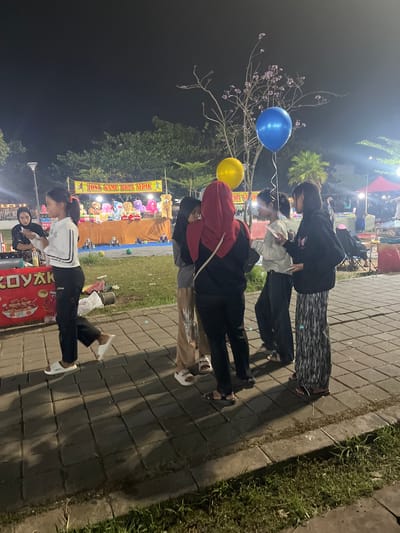 At night in Sodong, Indonesia, several young individuals are gathered near a game booth. A girl in a white shirt and black pants walks past a food stall. In the background, a banner reads "ZONA GAME BOLA SEPAK," indicating a soccer ball game. There are stuffed animals on display at the game booth, and several people are standing around, some looking at their phones. Two balloons, one blue and one yellow, float in the air. The scene appears to be an outdoor market or festival, with tents and lights visible in the distance.