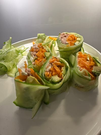 The image displays a white plate filled with six cucumber-wrapped food rolls and additional cucumber ribbons, situated in an indoor setting within the city of Latsia, Cyprus. Each roll is constructed from thin, pale green cucumber slices forming the exterior, encasing a filling composed of shredded orange carrots, light pinkish-brown chunks of tuna, creamy white dressing, and slices of green avocado. One of the rolls has visible shredded carrot strips on the plate next to it. The rolls are arranged loosely on the plate, with some standing upright and others leaning. The plate rests on a subtly textured, light-colored surface, possibly a countertop. The background is a plain, light grey wall or surface. No individuals are present.