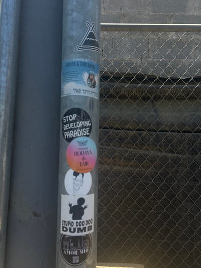 The image shows a metal pole covered in stickers located in the Cobano District, Costa Rica. The pole is a street light, with the stickered side facing a chain-link fence. The fence is in front of a gray concrete wall with a black object, possibly a tarp or plastic sheeting, lying on top. Several stickers cover the pole. The topmost is a black and white drawing of a tent with a mountain peak, followed by a sticker of two people in a photo with text in Hebrew. Below the photo is a sticker that reads "STOP DEVELOPING PARADISE". Under that sticker is a pink circular sticker that reads "CONSTRUCTOR HODAYA & TAIR" and a sticker of a man's head, facing forward, in black and white. The bottommost sticker reads "STUPID DOO DOO DUMB" in black text inside of a black circle with white text.