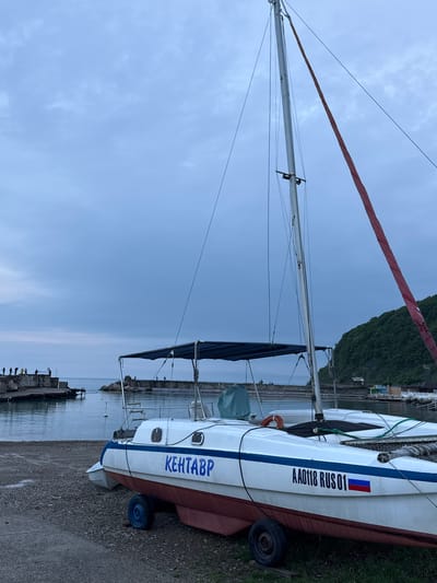A sideways outdoor shot features a sailboat on a trailer next to a body of water under a cloudy sky. The sailboat is white with a red waterline and a blue stripe along the side. The name "КЕНТАВР" is written vertically in blue on the side of the boat. Above this, "AA0118 RUS 01" is printed vertically in black, with a small Russian flag. The mast is tall and silver. The boat sits on a black-wheeled trailer on a gravelly surface near a grassy area.

In the background, a rocky shoreline and a pier are visible along the water's edge, where several people are fishing. The background includes a green, tree-covered area. The sky is overcast, indicating a potentially gloomy or overcast day. This scene is situated in or near Novomikhaylovsky, Russia, as evidenced by the Cyrillic lettering and "RUS" marking on the boat.