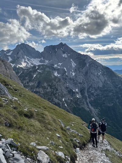 The image depicts a mountainous landscape with a clear sky and scattered clouds. In the foreground, a narrow, rocky path winds through a grassy slope. Three individuals are walking along the path, all facing away from the camera. The person in the middle is wearing a white cap and a backpack with an orange reflector. The person to their right is wearing a hat and a backpack. They are all equipped with trekking poles. The middle ground is dominated by rugged, rocky mountains, some of which are partially covered in snow. The background shows more distant, layered mountain ranges and a dense forest. The lighting suggests it is daytime, with sunlight illuminating the scene. This scene is located in the vicinity of Jošanica, Montenegro, a region known for its dramatic mountain terrain and hiking opportunities.