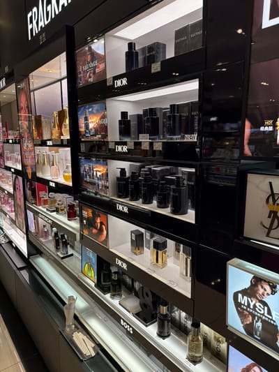 The image depicts a well-lit, modern retail display of fragrances in a store located in Nanjing Shi, China. The display features multiple shelves showcasing various perfume bottles and their packaging, predominantly from brands like Dior, Sauvage, and Boss. The items are arranged neatly, with product boxes and individual bottles clearly visible. Some shelves have promotional images or advertisements, including a picture of a man with the text "MYSLF" and another featuring a model with "SAUVAGE" branding. The overall environment suggests a high-end department store or a specialized fragrance boutique, emphasizing a clean and organized presentation to attract customers. There are no people visible in the frame.