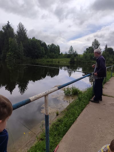 The image shows an outdoor scene in Советско-Никольское, Russia, by a body of water. An older man with gray hair stands on a concrete path, facing the water and holding a fishing rod. He is wearing a dark blue jacket with a striped shirt underneath and dark pants. He appears to be smoking. In the foreground, a child's head and shoulder are visible, looking towards the water. Another child, with a yellow shirt, is partially visible in the bottom right corner. The water is calm and reflects the surrounding trees and cloudy sky. There is a metal railing with blue and white paint separating the path from the water. Lush green grass grows along the edge of the water and the path.