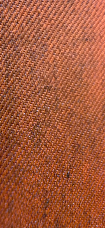 The image is a close-up, angled shot of an orange fabric. The fabric has a visible woven texture with threads running diagonally. There are some small dark specks and possibly some lint on the surface of the fabric. The lighting appears to be coming from the right side, casting subtle shadows and highlighting the texture. There is no indication of a specific location in Hatting, Denmark, or any people or actions. The image is purely a detailed view of a textile material.