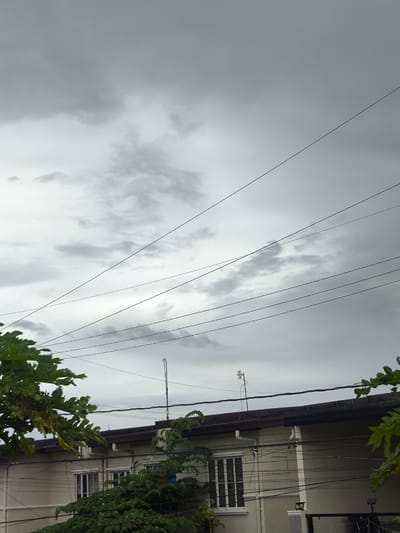 A low-angle, medium shot captures a residential building under a cloudy sky, as seen from General Trias, Philippines. The building's facade is light-colored, with white-framed windows. Several dark electrical wires crisscross the frame, suspended above the building and a lush green tree in the foreground. The sky is overcast with grey clouds, indicating a potentially rainy day. Two antennas are visible on the rooftop of the building.