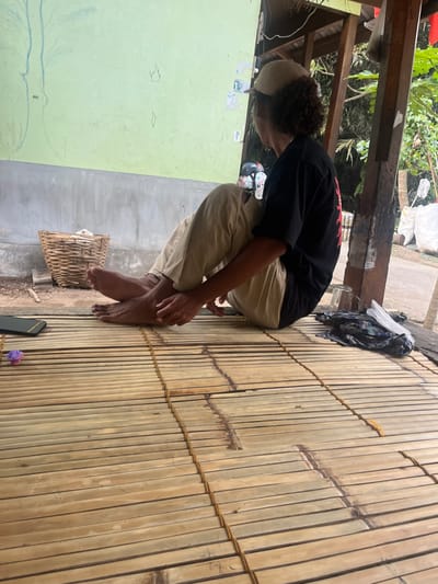 The scene depicts an individual seated on a light-colored bamboo slat platform in Desa Batu Layar, Indonesia. The person, appearing male, wears a black short-sleeved t-shirt with a red design visible on the back, light-colored long pants, and a tan baseball cap worn backward over dark, curly hair. The individual is barefoot, positioned with knees bent and feet flat on the platform, facing away from the camera and oriented towards a light green painted wall with a gray concrete base. Faint blue lines and small, light-colored paper fragments are visible on the green wall.
On the bamboo platform to the left of the seated individual, a black smartphone with a yellow case rests horizontally, adjacent to a small purple, flower-like object. On the ground next to the concrete wall base, a woven bamboo basket contains white material. To the right of the individual, a clear glass mug and a crumpled dark fabric item are present on the bamboo platform. The immediate background features brown wooden structural beams, and beyond, green foliage is visible, indicating an outdoor or semi-open environment. A dirt path or ground is discernible to the right of the wooden beams.