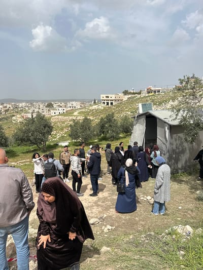 The image depicts an outdoor scene in Dura, Palestinian Territory, on a partly cloudy day. In the foreground, a woman in a dark brown abaya and hijab walks from left to right, partially obscured. A man in a grey shirt and blue jeans stands to her left, facing away. Red and white striped tape is visible on the ground near his feet.
The midground features a gathering of approximately 20-30 individuals, predominantly adults, on an unpaved, uneven dirt path with sparse green vegetation and rocks. The group comprises men in casual clothing (jeans, jackets, shirts) and women, many of whom wear hijabs and long modest clothing (abayas, dresses) in various colors, including dark hues, blue, and grey. Individuals are standing in small clusters, some appearing to be engaged in conversation or observing a point of interest. One woman in a blue abaya and black hijab holds a smartphone, appearing to photograph or record.
To the right midground, a grey, temporary or makeshift structure with a rectangular opening is visible. A small solar panel is mounted on its roof. Scattered olive trees with green foliage are present across the hillside behind the gathering.
The background reveals a panoramic view of the city of Dura, extending across a valley and up several hillsides. Numerous light-colored, multi-story buildings, some appearing unfinished, are visible. Patches of green vegetation are interspersed among the structures and across the hilly terrain. Distant hills mark the horizon under a sky with scattered white clouds.