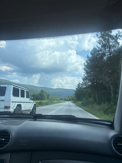 The image was taken from inside a car on a sunny day, looking out through the windshield. A white Mercedes-Benz G-Class is visible in the left lane of a paved road. The road stretches into the distance, lined with trees on either side. Rolling hills covered in green foliage are visible in the background under a partly cloudy sky. The car's dashboard and windshield wipers are in the foreground. The scene is consistent with a rural road in Leshnitsa, Bulgaria, given the mountainous terrain and vegetation. There are no people clearly visible in the image.