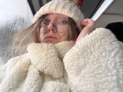 The image presents a close-up, slightly upward-angled view of a single individual, likely female, situated indoors or within a vehicle. The specific location within Борисово, Russia, cannot be determined from the visual evidence. The individual is wearing a cream-colored, ribbed knit beanie hat and a high-collared, textured cream-colored fleece jacket. Round, thin-framed eyeglasses are worn, and strands of light-colored hair are visible, some extending across the left side of the frame. The individual's gaze is directed upwards and to the right. A hand is positioned near the right side of the head, appearing to rest near the hat. To the left, a window pane is partially visible, displaying condensation or moisture droplets. Through this window, the exterior appears as a blurred, indistinct grey environment. In the upper right background, a partial view of a red object with white lettering or graphics is discernible. The immediate background on the right is dark and obscured.