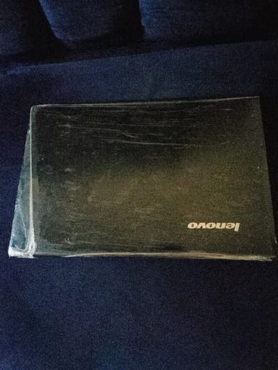 This image, reportedly taken in Okinni, Nigeria, presents a black Lenovo brand laptop resting on a dark blue, textured textile surface. The closed laptop is partially enveloped in a clear, wrinkled plastic film that covers the lid. The white "Lenovo" logo is distinctly visible on the lower right quadrant of the lid, appearing inverted relative to the top of the image frame. Surface wear, including scratches and smudges, is apparent on the laptop lid beneath the plastic film. The background consists of a uniformly dark blue textile material. No individuals or active interactions are depicted.