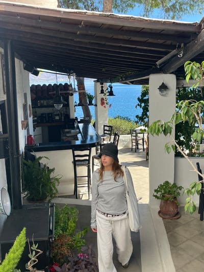 The image displays an outdoor dining area of a restaurant or cafe situated in Megalochori, Greece, with a clear view of a large body of deep blue water and distant landmass under a bright blue sky.

In the foreground and midground, a single female subject with shoulder-length light brown hair is visible, wearing a dark grey bucket hat, a light grey long-sleeved top, white wide-leg trousers, and carrying a white tote bag on her left shoulder. She is walking towards the right side of the frame, looking slightly to her left.

The establishment features white stucco walls and a dark wooden pergola roof structure providing shade. To the left, a black, curved bar counter with three high-backed bar stools is present. Behind the bar, shelves hold a red kettle, a coffee maker, and various other containers. A disco ball and other hanging light fixtures are suspended from the wooden beams. In the lower left foreground, a black, square-shaped oven or stove is visible, surrounded by potted plants including ferns and flowering species. To the right of the woman, and further into the background, a paved patio with several dark wooden chairs and light-colored tables is arranged, offering seating with a direct view of the sea. Additional potted plants and a large tree trunk are also visible on the right side of the frame. The scene is illuminated by natural daylight, casting shadows.