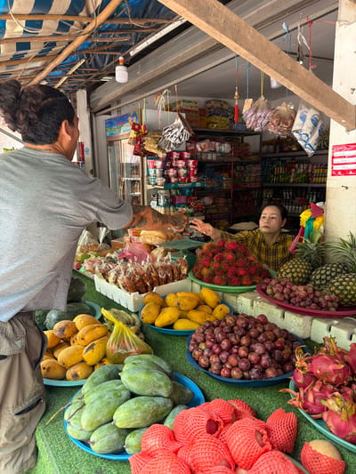 In Xieng Men cultural village, Laos, a vendor is handing fruit to a customer at an outdoor stall. The customer, a man with his back to the camera, wears a grey t-shirt and has his hair tied up. The vendor, a woman in a yellow plaid shirt, reaches out from behind a display of rambutans and grapes. The stall is filled with a variety of fresh produce, including pineapples, mangoes, dragon fruit, and other items. Shelves stocked with packaged goods are visible in the background, under a striped tarp roof supported by bamboo.