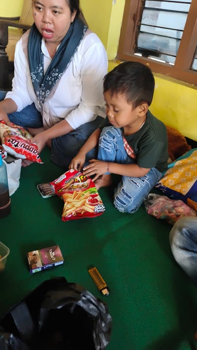 In a room with yellow walls and a brown window frame, a woman and a young boy are sitting on a green carpet. The woman, wearing a white shirt and jeans, is holding a bag of snacks. The boy, in a dark green t-shirt and ripped jeans, is looking at another bag of snacks, which appears to be french fries. On the carpet, there are also a pack of Camel cigarettes, a yellow lighter, and a black plastic bag. The room is lit by natural light coming from the window.