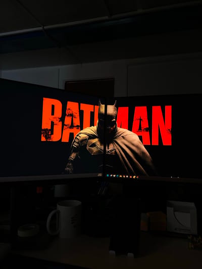 The image shows a dimly lit office desk with two monitors. The left monitor displays a portion of the movie poster for "The Batman," featuring the character's silhouette and the movie title in large, distressed orange lettering. The right monitor shows the other half of the poster, with the "MAN" part of the title and more of Batman's cape and cowl. The monitors are angled together, creating a wide-screen display.

On the desk in front of the monitors, several objects are visible. To the left, there is a white mug with a logo that appears to be "RBM" or "REN," and a roll of tape. In the center, a smartphone is propped up on a stand, and behind it, there are stacks of colorful sticky notes and a white tissue box with a pattern. To the right, there is a small container with colorful items that could be pens or markers.

The overall lighting suggests it is nighttime or the office lights are turned down. The background above the monitors appears to be a ceiling with fluorescent lighting fixtures, typical of an office environment. There is no clear indication of the location in the city of Yukhta, Russia.