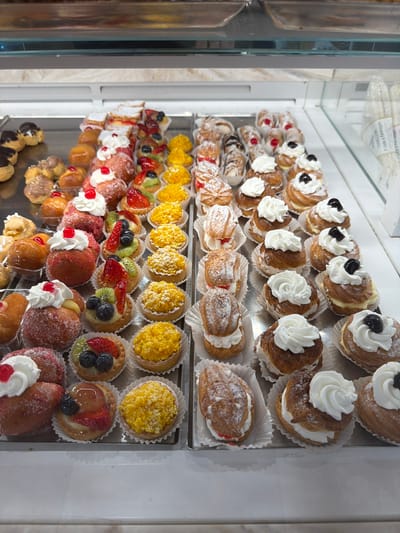The image depicts a refrigerated display case filled with Italian pastries, most likely in a bakery or pastry shop. The location is likely in Tarquinia, Italy, given the traditional Italian pastries on display. The display case has multiple shelves with pastries arranged in rows.

The top shelf holds cream puffs, some topped with whipped cream and black cherries. The second shelf contains pastries dusted with powdered sugar, with a cream filling. The third shelf features small tarts filled with yellow cream or custard, and the fourth shelf features fruit tarts with sliced strawberries, kiwi, and blueberries in a clear glaze. The bottom shelf showcases a variety of pastries topped with whipped cream and cherries. The scene suggests a typical Italian confectionery establishment.