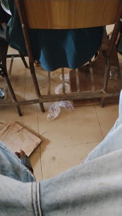 A low-angle shot captures a scene underneath a chair. The chair has a wooden back and a dark cushion. Litter, including plastic bags and a piece of cardboard, is scattered on the beige tiled floor. The bottom portion of the image shows the lap of a person wearing blue jeans. The overall image is somewhat cluttered and unfocused.