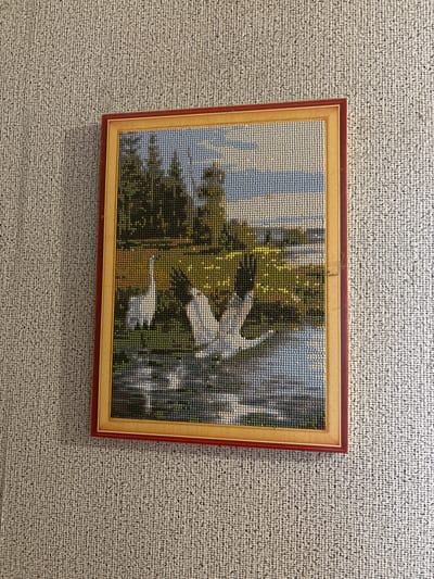 The image shows a framed diamond painting hanging on a textured wall. The diamond painting depicts a landscape scene with two swans in a body of water. The sky is depicted with blue and white squares, and the trees and grass are rendered in various shades of green and yellow. The water reflects the sky and the surrounding greenery. The frame is a double frame, with an inner gold-colored frame and an outer dark red frame. The wall appears to be covered in a light-colored, woven material. The diamond painting is centrally located in the frame and appears to be the sole subject of the image. There are no people or animals other than the swans in the painting. The image does not provide any information about the specific location within the city of Ильпанур, Russia, or any details about the context of the painting's display.