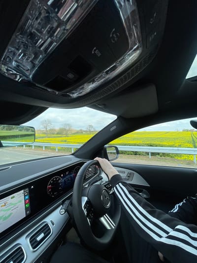 The image shows the interior of a car, from the driver's perspective. The driver is holding the steering wheel with their right hand and their left arm is resting on the seat. The driver is wearing a black jacket with white stripes. The dashboard shows a digital display with a speedometer, a temperature gauge, and a navigation system.  The car is traveling on a road with a yellow field on the right side. There is a metal fence separating the road from the field.  The car is driving towards a row of trees in the distance.  The GPS shows a location that is in close proximity to the city of Woburn, United Kingdom, which is located at a latitude of 51.99 and a longitude of -0.618. The coordinates provided indicate a rural setting, likely in a field area outside of the main town center.  The sky is cloudy.  The car's interior is illuminated by natural light.  The overhead light is off.  The driver's side window is down, and the interior mirror is tilted.  The dashboard is displaying a number of 37. The rearview mirror is visible.  The steering wheel is leather-wrapped with a silver emblem.  The buttons on the overhead console indicate an SOS function, a microphone, and a button labelled 'SOS'.  The door handle has a button.  The seat is covered in a dark fabric.  The car has a black interior.