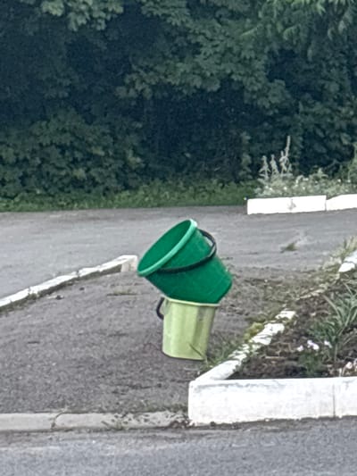 Two plastic buckets, one green and one lime green, are stacked on an asphalt surface. The green bucket is on top, inverted and slightly askew, with its rim resting on the bottom bucket. The lime green bucket is upright. Behind the buckets, there is a strip of green vegetation, a concrete border, and more asphalt. The scene is outdoors, likely in a residential or public area. The image does not contain any information that would allow for the identification of the location as Gukovo, Russia.