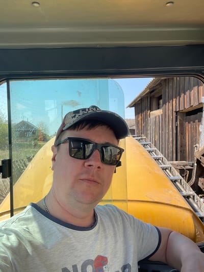 A man wearing a baseball cap and sunglasses is pictured inside the cab of a yellow construction vehicle in Поедуги, Russia. The man is wearing a gray t-shirt and a silver chain. Outside the window of the cab, there is a partially visible wooden building, and there is equipment visible above the man.