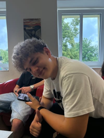 The image, taken in Löcknitz, Germany, shows two young men indoors during daytime. The perspective is tilted, with the original image rotated 90 degrees clockwise.

In the foreground, a young man with curly brown hair, wearing a white t-shirt with "NIKE" partially visible, looks towards the viewer with a slight smile. His left hand, wearing a black wristband, points at a smartphone held by another person.

The second individual, partially visible, wears a dark t-shirt with some illegible white text and light blue denim shorts. They are seated on a red couch or chair, holding the smartphone being pointed at. A silver-toned watch is visible on their left wrist. The activity appears to be a casual interaction, with one person showing or sharing something on a smartphone screen with the other or the viewer. The phone screen displays a blurry image, possibly of an interior space.

In the background, a large window reveals a bright blue sky with some clouds and lush green trees, indicating pleasant weather. To the left of the window, a framed picture resembling Edvard Munch's "The Scream" hangs on a white wall. The room seems to be a casual living space or common area. The bright outdoor light suggests it is daytime. The overall mood is relaxed and friendly.