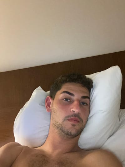 An adult male with curly dark hair, blue eyes, and light facial stubble lies shirtless on his back, propped against a white pillow. He looks directly forward. A dark brown headboard or wall panel is visible behind the pillow, with a light-colored ceiling above. The scene appears to be an indoor setting, likely a bedroom or hotel room, located in the City of Albany, United States.