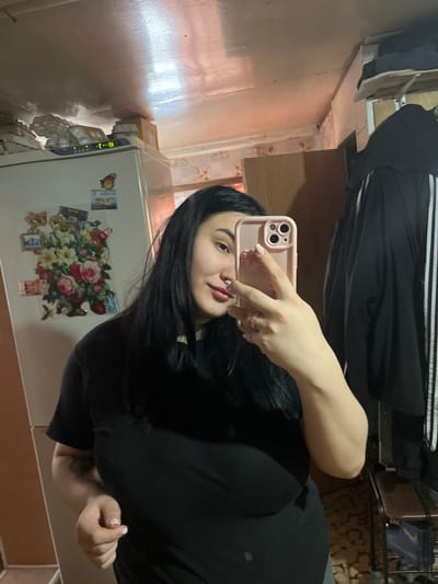A female individual with long, dark hair and a light complexion, wearing a black t-shirt, is depicted taking a mirror selfie in an interior setting located in Романята, Russia. She holds a light pink smartphone with a matching case in her right hand, which displays a light pink manicure and a ring on her ring finger. Her left arm exhibits a partial tattoo. The background to her left features a white refrigerator adorned with multiple magnets, including a prominent floral design, and smaller rectangular magnets. On top of the refrigerator, several white egg cartons are present, along with a digital display showing "18:81". The wall behind the refrigerator has a patterned wallpaper. To her right, a black jacket with three white stripes, characteristic of the Adidas brand, hangs on a visible rack or hook. Above this, a shelf with dark objects is visible. A portion of a chair with a brown upholstered seat is also discernible in the lower right. The ceiling is light-colored and reflective, while the flooring appears to be a patterned surface.