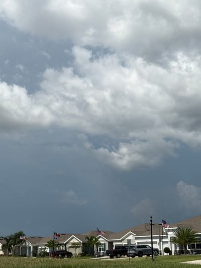 In Parrish, Florida, a row of modern houses with gray roofs and white trim is visible under a cloudy sky. The sky is filled with dramatic, dark clouds, suggesting an impending storm, with lighter clouds scattered throughout. In front of the houses, there is a grassy area and a paved driveway where several vehicles are parked, including a red SUV, a black pickup truck, and a silver SUV. American flags are displayed on flagpoles in front of some of the houses, and palm trees are scattered around the properties. The overall scene conveys a sense of suburban life under a potentially tumultuous sky.