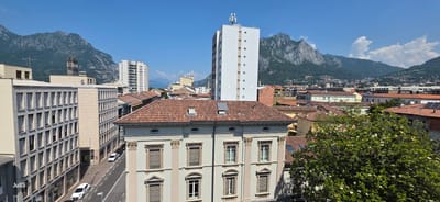 This is an eye-level, medium shot, outdoor, daylight, horizontal photograph of the city of Lecco, Italy, captured on a sunny day. The image depicts a cityscape with a foreground featuring a light-colored building with classical architectural elements, including columns and decorative window surrounds, and a red-tiled roof. To the left of this building, there is a modern, multi-story building with a grid of windows. A street runs along the side of these buildings, with two white cars visible. In the middle ground, more red-tiled roofs of various buildings are visible, interspersed with a few modern structures, including a tall white building with antennae on its roof and another white high-rise building. The background is dominated by the dramatic, green, and rocky mountains characteristic of the Lecco region, under a clear blue sky with scattered white clouds. The overall scene conveys a sense of urban life nestled within a mountainous landscape. The text "JullS" is visible in the lower-left corner of the image.