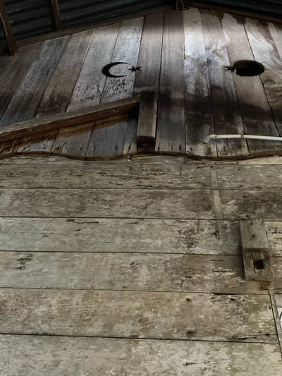 The image depicts the interior of a rustic wooden structure, likely a barn or storage shed, in Blangkabu, Indonesia. The walls are constructed of weathered, vertical wooden planks, showing signs of age and wear, with some discoloration and peeling paint. A corrugated metal roof is visible at the top.

Prominently displayed on the upper section of the wall is a cut-out of a crescent moon and star, a symbol often associated with Islam. To the right of this symbol, there is a circular hole in the wooden wall, appearing to be a small window or opening. A horizontal white pipe runs across the wall above a lower section of planks.

The lower portion of the image shows more of the weathered wooden wall, with horizontal planks that are heavily textured and show signs of decay. A wooden fixture, possibly a latch or support, is attached to the wall on the right side. The overall impression is one of a functional, utilitarian space that has been exposed to the elements for a considerable time. There are no people or animals visible in the frame.