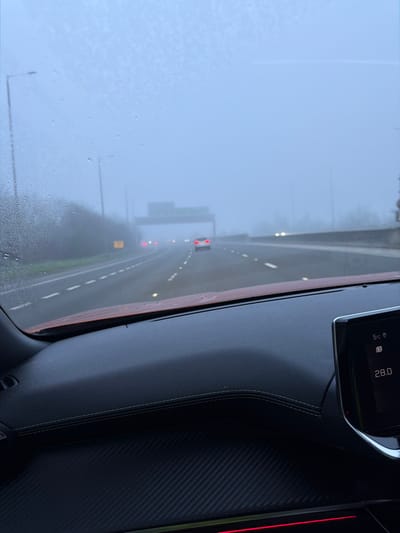 The image depicts a multi-lane motorway scene observed from the interior of a vehicle, through a windshield partially obscured by water droplets. Heavy fog significantly reduces visibility. The dashboard in the foreground features dark, textured surfaces, including a section with a carbon fiber pattern and another with white stitching. A digital display on the right shows "5°C" and "28.0." On the motorway, multiple vehicles are visible ahead, indicated primarily by their illuminated red rear lights and white headlights piercing the fog. A white passenger car is discernible in the middle lane further down the road. Streetlights line the left side of the motorway, and a large overhead gantry-style road sign structure is faintly visible in the distance. The scene is located on a motorway in or near Garden City, United Kingdom. No individuals are visible.
