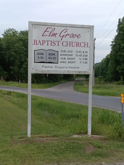 The image shows a sign for Elm Grove Baptist Church located on a roadside. The sign provides the church's name in a decorative red font at the top, followed by "BAPTIST CHURCH." Beneath this, there are Bible verses "JOHN 3:16" and "ROM. 10:13" presented next to each other. Further down are the service timings: "SUN. SCH 9:45 A.M.", "WORSHIP 10:45 A.M.", "SUN. NIGHT 5: P.M.", and "WED. NIGHT 7: P.M." The sign also identifies the pastor as "Eugene Kester." The sign is supported by two white posts. The background features a road leading uphill into a wooded area. Green vegetation covers the ground around the sign. There is another road sign on the right. The location appears to be rural and likely in the United States, potentially near the city of Stilwell.