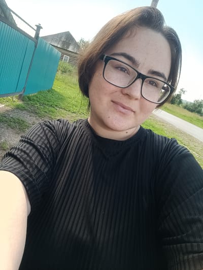 A young woman with dark hair and glasses is taking a selfie outdoors. She is wearing a black, ribbed t-shirt. Behind her, there is a turquoise metal fence, a wooden house with a slate roof, green grass, and a dirt road. The sky is blue with some haze. The scene appears to be in a rural area, possibly on the outskirts of Усть-Тунтор, Russia, given the presence of a traditional wooden house and fence.