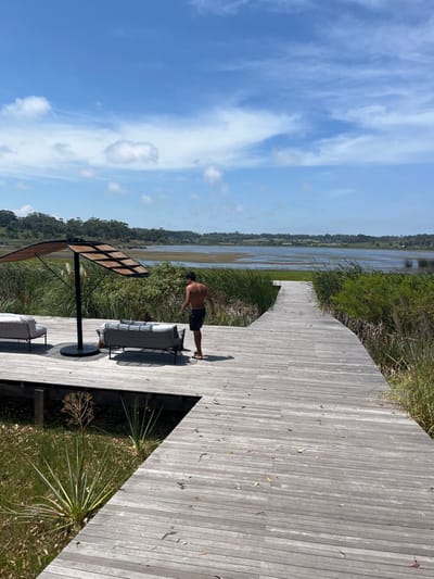 A person stands on a wooden boardwalk beside a tranquil body of water, possibly a lake or a bayou, under a bright blue sky. Lush green vegetation lines the opposite bank. Nearby, a patio set with a sun umbrella offers a relaxing space. The boardwalk extends into the distance, inviting exploration along the scenic waterfront.