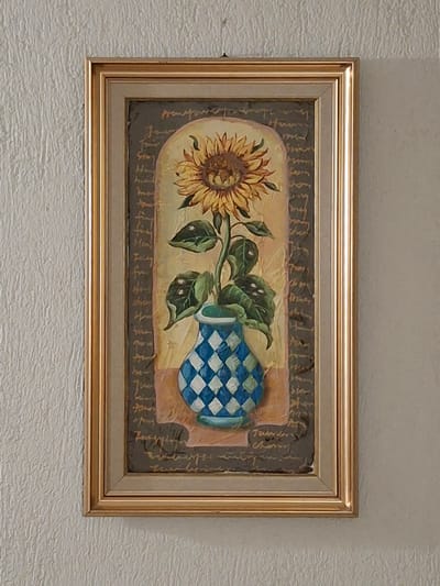 The image shows a framed painting of a sunflower, leaves, and a blue-and-white checkered vase. The painting is rectangular and has a gold frame. The background of the painting is a light beige color with a dark brown border. The painting depicts the sunflower and leaves extending to the left and the vase to the right, the scene appears to be of an interior. There is no information about the location in Krumovgrad, Bulgaria.