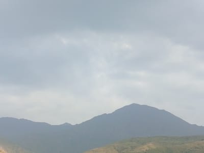 The image depicts a misty mountainous landscape under an overcast sky. In the foreground, rolling hills with sparse vegetation are visible. The middle ground is dominated by the silhouetted forms of several mountain ranges, their peaks softened by the atmospheric haze. The sky above is a uniform, muted gray, with faint cloud formations suggesting a diffused light source. The overall scene is one of serene, natural grandeur, typical of highland environments. This specific view is located in the vicinity of Jardy-Su, Kyrgyzstan, a region characterized by its vast mountainous terrain. There are no people, animals, or man-made structures visible in the frame.
