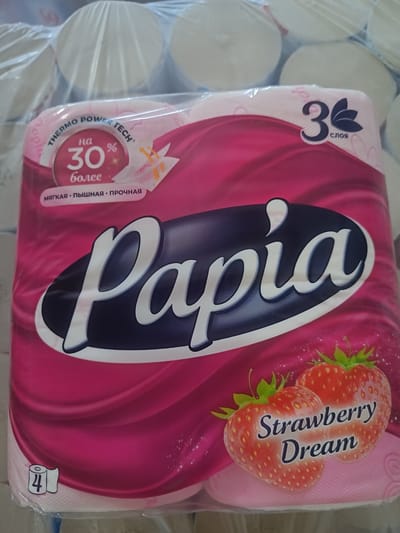 A plastic-wrapped package of Papia brand toilet paper is shown. The packaging is primarily pink and features swirling patterns. The brand name "Papia" is prominently displayed in large, white, stylized letters on a dark blue oval. Above the brand name, a circular graphic indicates "на 30% более" (for 30% more), with "THERMO POWER TECH" written around the edge. To the right of this graphic, a numeral "3" is shown with a stylized leaf design below it, indicating "3 слоя" (3 ply). Two red strawberries with green leaves are depicted at the bottom right, with the words "Strawberry Dream" written in white text over them. A small icon of a roll of toilet paper with the number "4" is visible in the lower left corner, suggesting it is a pack of 4 rolls. The background consists of stacks of white toilet paper rolls wrapped in plastic. The scene is a close-up, frontal view of the product packaging. There is no indication of a specific location in the city of Berdiansk, Ukraine, or any people.