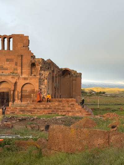 The image depicts the ruins of Ani Cathedral in Anipemza, Armenia. The structure is predominantly brown stone with arched openings. Three people are visible: one dressed in orange on the steps, one in yellow above them, and one in black on the top of the wall, all situated within the ruins. A landscape with hills and sky are visible in the background. Grass and some debris are present on the ground surrounding the structure.