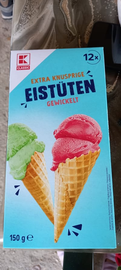 The image shows the packaging for a box of 12 ice cream cones. The box is light blue with text in German. The front of the box features two ice cream cones with scoops of ice cream, one green and one red. The text on the box reads "CLASSIC EXTRA KNUSPRIGE EISTÜTEN GEWICKELT" and "12x". There is also text on the right side of the box that appears to be in multiple languages and is too small to read clearly. The bottom left corner of the box shows "150 ge". The background of the image is a blurry surface that appears to be a table with a marbled pattern. The image is a product shot of the ice cream box. There is no indication of the city of Iskra, Bulgaria in this image.