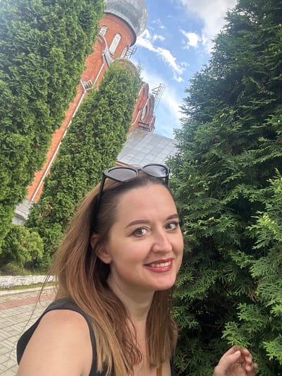 In Medvedevo, Russia, a woman with shoulder-length brown hair and sunglasses perched on her head takes a selfie outdoors. She is smiling and wearing a black top. To her left, a red brick building with a silver dome is visible behind tall, green evergreen trees. To her right, a dense wall of evergreen foliage fills the frame. The background shows a blue sky with white clouds. The ground in front of her appears to be paved with interlocking stones.
