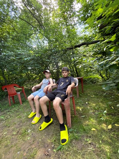 Two young boys, appearing to be around 8-10 years old, are seated on plastic chairs in an outdoor, wooded area. The boy on the left wears a gray tank top, denim shorts, a red and white baseball cap, and yellow slide sandals. The boy on the right is dressed in a dark blue t-shirt with a gold number "7" and emblem, black shorts, a dark baseball cap, and black socks with yellow slide sandals. They are positioned in front of a red table. The ground is a mix of dirt and grass, with fallen leaves scattered about. Lush green foliage and trees form the backdrop, suggesting a natural, possibly secluded, setting. The lighting indicates it is daytime, with sunlight filtering through the leaves. The location is identified as Аряж, Russia.