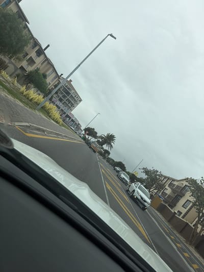 The image depicts a street scene in Blouberg, South Africa, viewed from inside a moving vehicle. The camera angle is tilted, showing the road and sidewalk at an angle. On the right side of the road are several vehicles, with a white Renault prominently visible. Further along the road is a McDonald's restaurant. The left side of the road features apartment buildings, a sidewalk, and a curb marked with yellow. A tall lamppost stands on the sidewalk. The sky is overcast.