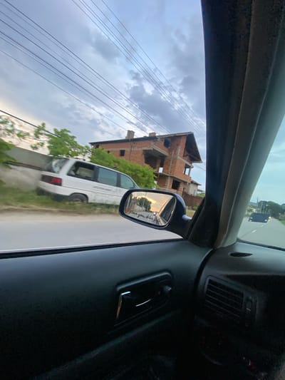 This image, taken from inside a car, captures a scene on a street in Miriantsi, Bulgaria. The sky is overcast with scattered clouds, and power lines stretch across the frame. In the background, a partially constructed brick building with multiple stories and exposed concrete elements stands prominently. To the left of the building, a white van is parked on a grassy area next to a low concrete wall. The van appears to be older and is partially obscured by trees and foliage. A side-view mirror of the car reflects a street scene with a sunset sky and headlights from an approaching vehicle. The interior of the car is visible in the foreground, showing part of the dashboard, a door handle, and an air vent. The road is paved and appears to be a residential street.