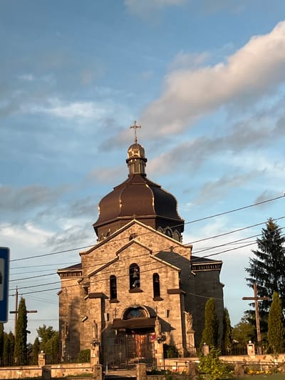 The image depicts a stone church located in Hrabovets, Ukraine, under a partially cloudy blue sky. The church features a roughly hewn light-colored stone facade with a gabled section and arched window openings, one of which visibly contains a bell. Several stone statues are integrated into the facade and at the base of the structure. A prominent dark, possibly metal, dome with a ribbed pattern crowns the main body of the church, topped by a smaller, reflective golden cupola. A golden Christian cross stands at the apex of the cupola. The front of the church and the golden cupola are illuminated by warm, direct sunlight, casting some shadows on the stone work. In the foreground, a low stone fence with decorative metal sections and small crosses runs horizontally across the frame. Beyond the fence, various coniferous trees, including tall, slender cypresses and a larger, darker evergreen on the right, are visible. Two large wooden crosses are situated behind the fence on either side of the church's immediate vicinity. Multiple black utility wires traverse the upper and middle portions of the scene. A partial blue rectangular sign is visible on the far left. No individuals are present in the image.