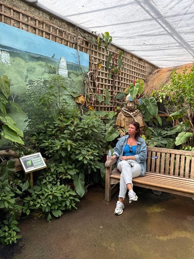 The image shows a woman reclining on a wooden bench in an indoor garden. She is wearing a blue top, denim jacket, light-colored jeans, and white sneakers. She holds a water bottle in her hand. A stone sculpture of a face with a headdress is positioned beside her. The bench is surrounded by lush greenery. Behind the vegetation, there is a lattice wall with a mural of a landscape painting. To the right of the painting, a sign with text and a leaf drawing is set among the greenery. A white, possibly translucent, covering is visible on the ceiling and upper left of the frame. The setting appears to be indoors and possibly a greenhouse or conservatory-like structure.

The location is Stratford-on-Avon, United Kingdom.