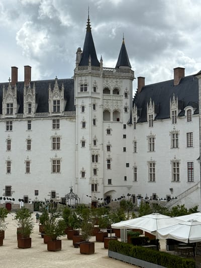 This image depicts the Château des ducs de Bretagne in Nantes, France. The imposing white stone castle features dark, pointed roofs and multiple towers, with a central tall tower dominating the skyline. Ornate architectural details, including decorative window frames and battlements, are visible on the castle's facade. In the foreground, a courtyard is furnished with several large white umbrellas providing shade over outdoor seating areas. Rows of potted plants, some appearing to be young trees, are arranged in weathered metal planters. A person is visible in the middle ground walking across the courtyard. The sky is overcast with visible clouds, suggesting a temperate climate.